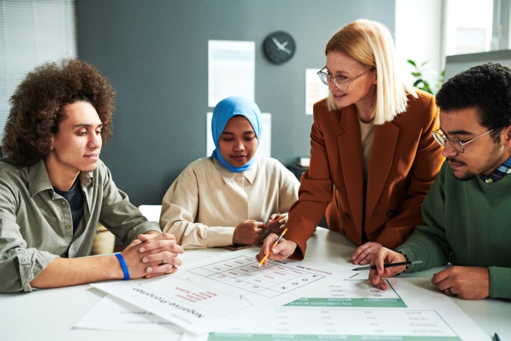 Tutor in glasses pointing to a worksheet while three students around the table look on