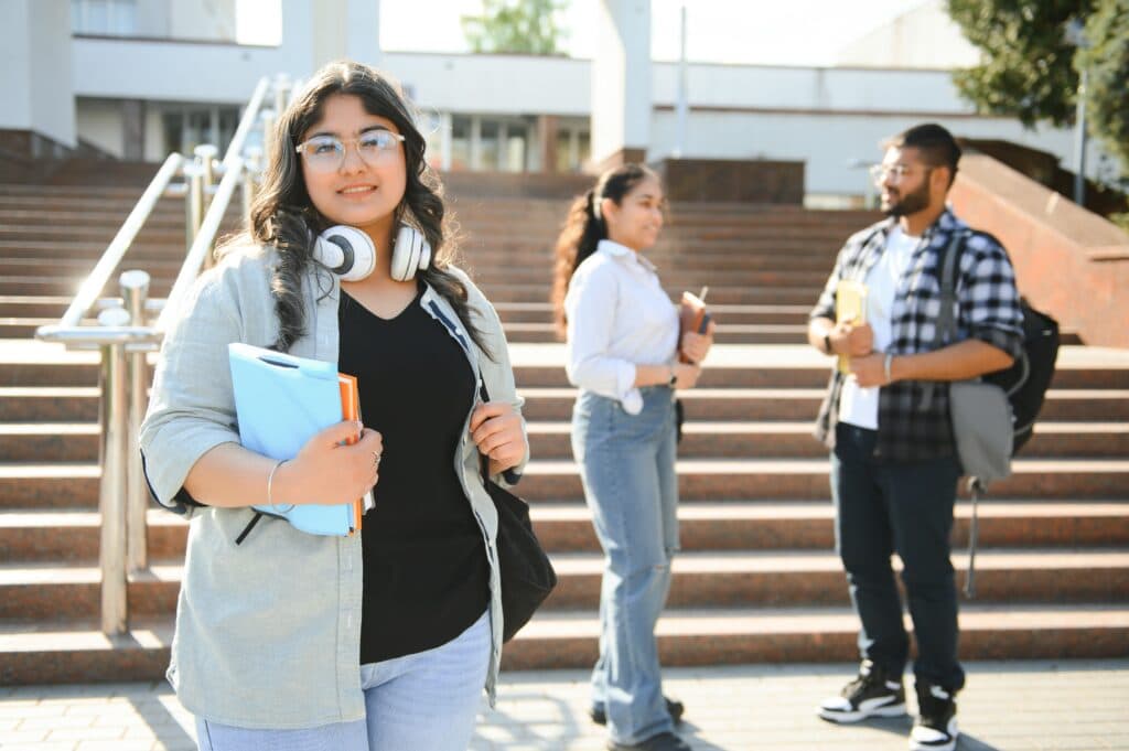 Smiling student holding folders on campus steps, with two classmates talking behind her